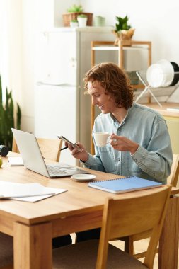 Yakışıklı genç adam düzenli bir mutfak masasında oturuyor, kahve, dizüstü bilgisayar ve telefonla birden fazla iş yapıyor..