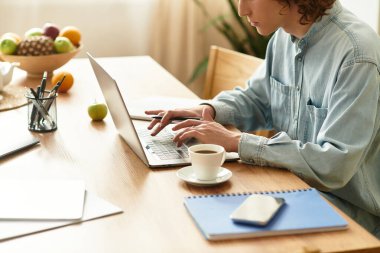 Odaklanmış genç bir adam ahşap bir masada oturur, dizüstü bilgisayarında yazar ve kahvesini yudumlar..
