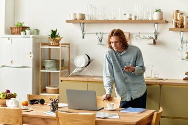 Yakışıklı bir genç adam, mutfak masasında düşünceli bir şekilde duruyor ve yakınlarda dizüstü bilgisayarı olan görevlere odaklanıyor.