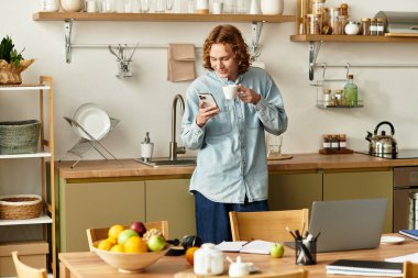 Sıcak bir mutfakta, genç bir adam bardaktan yudumlar ve telefonunu kullanır..