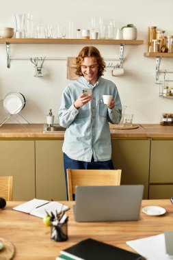 Yakışıklı bir genç adam rahat mutfağında duruyor, elinde bir bardak ve telefonuna sarılmış..