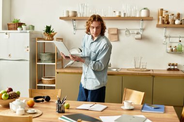 Yakışıklı bir genç adam parlak bir mutfakta duruyor, birden fazla iş yaparken dizüstü bilgisayarına odaklanıyor..