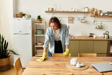 Kıvırcık saçlı, eldiven giyen genç bir adam şık bir mutfak ortamında ahşap bir masayı temizliyor..