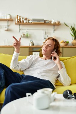 Yakışıklı bir genç adam açık sarı bir kanepede uzanıyor, telefonda mutlu bir şekilde sohbet ediyor..