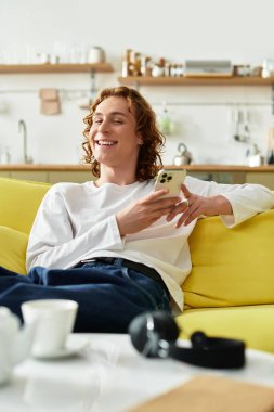 Neşeli genç bir adam evinde rahat bir şekilde uzanır, akıllı telefonunu yüzünde bir gülümsemeyle kullanır..