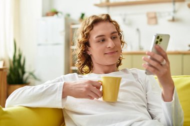 Sarı kanepede oturan genç bir adam kahvesini yudumluyor ve rahat bir evde telefonuna bakıyor..