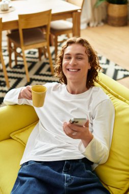 Yakışıklı genç adam evinde telefonuyla gülümseyen bir kupayla rahatça uzanır..