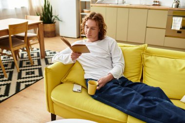 Yakışıklı genç bir adam sarı bir koltukta rahatça oturuyor, bir kitaba gömülmüş..