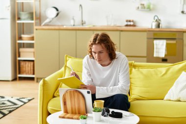 Çalışmaya dalmış genç bir adam, parlak ve davetkar bir yaşam alanında bir not defterine notlar alıyor.