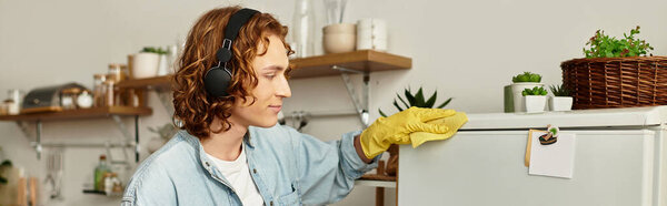 Счастливый молодой человек в повседневной одежде чистит холодильник в наушниках и жёлтых перчатках.