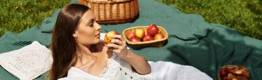 Genç bir kadın yeşil bir battaniyeye uzanıp parkta pikniğin ortasında taze bir elmanın tadını çıkarıyor..
