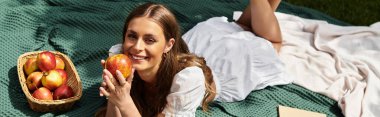 Genç bir kadın güneşli bir piknik sırasında etrafında taze elmalarla bir battaniyenin üzerinde uzanırken gülümsüyor..