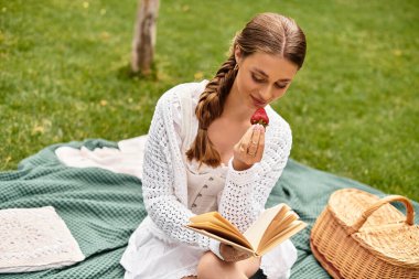 Genç bir kadın piknik battaniyesine oturmuş, yemyeşil bir parkta çilek okuyor ve eğleniyor..