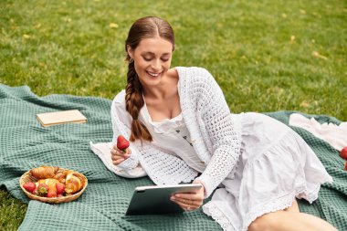 Genç bir kadın parkta bir battaniyeye uzanıyor, lezzetli atıştırmalıkların ve okumaların tadını çıkarıyor..