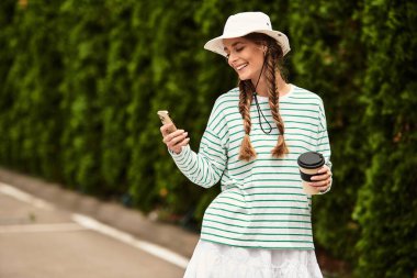 Saçı örgülü genç bir kadın parkta piknik yaparken telefonunu kontrol ederken gülümsüyor..