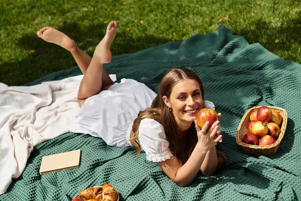Güneşli bir günde taze meyve ve hamur işleriyle çevrili pikniğin tadını çıkaran gülümseyen genç bir kadın.