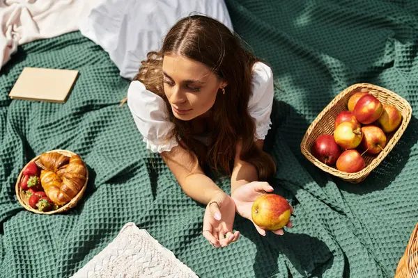 Genç ve güzel bir kadın piknik battaniyesinde dinleniyor, doğanın ortasında parlak bir elmanın tadını çıkarıyor..