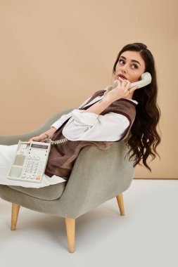 Uzun kahverengi saçlı genç bir kadın modaya uygun bir şekilde oturmuş, klasik bir telefonda sohbet ediyor..