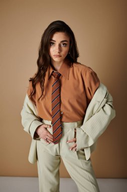 Stüdyoda, uzun kahverengi saçlı genç bir kadın kravat takan şık bir takım elbise giyiyor..