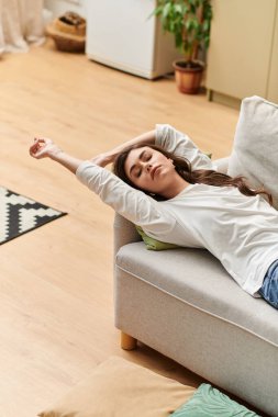 Kahverengi saçlı kadın rahat bir kanepede uzanırken günlük kıyafetlerini giyer.