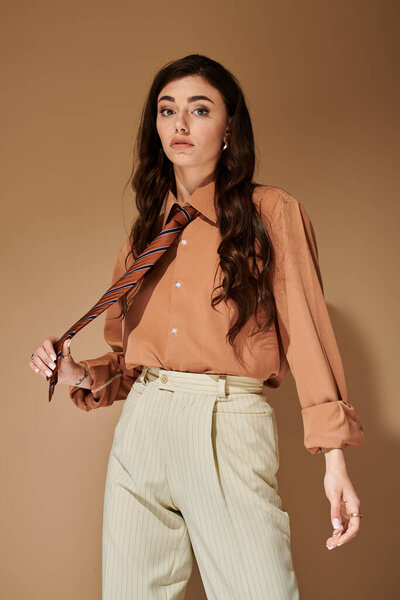 A stylish young woman with long brown hair poses confidently in a fashionable attire and tie.
