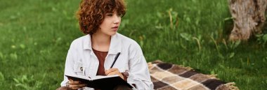 Kıvırcık saçlı genç bir kadın serin bir anın tadını çıkarıyor. Çimlerin ve afişlerin arasında bir battaniyeye yazı yazıyor.