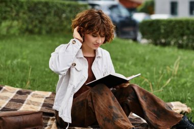 Kıvırcık saçlı genç bir kadın yeşil bir parkta, bir kitap okurken battaniyede dinleniyor..