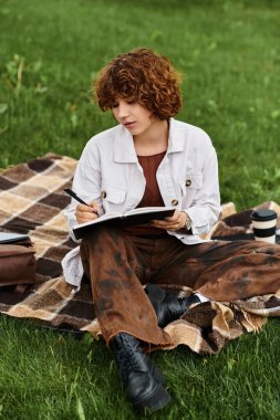 Kıvırcık saçlı genç bir kadın parkta güneşli bir günün tadını çıkarıyor, defterine fikirler çiziyor..