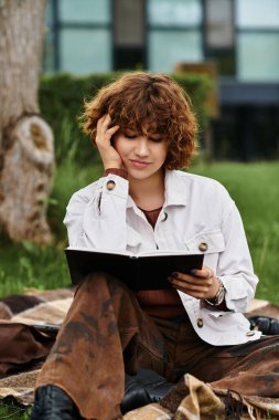 Kıvırcık saçlı genç bir kadın, canlı bir parkta battaniyede otururken kitap okumaktan zevk alır..