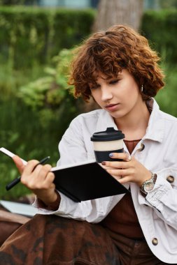 Kıvırcık saçlı genç bir kadın battaniyeye uzanır ve kahvesini içerken günlüğüne bir şeyler yazar..