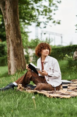 Kıvırcık saçlı genç bir kadın yeşil bir parkta battaniyeye sarılıyor, kitabına dalmış..
