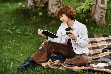 Kıvırcık saçlı kadın, parktaki defterine bir şeyler yazarken battaniyeye oturup kahvesini yudumluyor..
