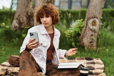 Kıvırcık saçlı kadın bir battaniyenin üzerinde uzanır, doğanın güzellikleriyle çevriliyken, telefonunu emer..