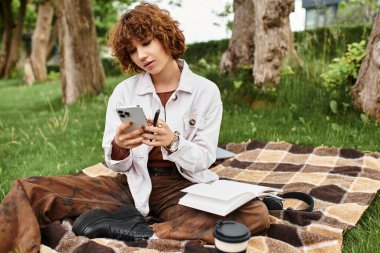 Yeşil bir parkta dinlenen kıvırcık saçlı genç bir kadın otururken telefonuyla meşgul oluyor..