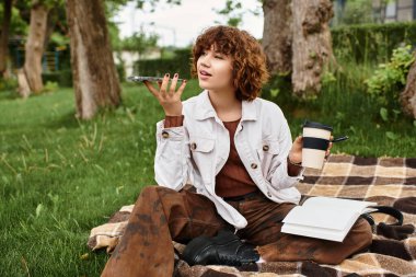 Kıvırcık saçlı kadın, yeşil bir parkta sohbet ederken battaniyeye oturup kahvenin tadını çıkarıyor..