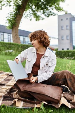 Kıvırcık saçlı genç bir kadın güneşli bir parkta dizüstü bilgisayarıyla uzaktan çalışıyor..