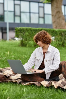 Kıvırcık saçlı genç bir kadın, parktaki dizüstü bilgisayarına odaklanmış bir şekilde battaniyeye oturur..