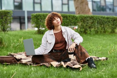 Kıvırcık saçlı kadın laptopunda çalışıyor, etrafı yeşil çimlerle çevrili, açık havada yaz atmosferini kucaklıyor..