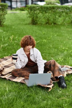 Kıvırcık saçlı genç kadın dışarıda kahvesini yudumlarken laptopuna odaklanmış bir şekilde battaniyeye oturuyor..