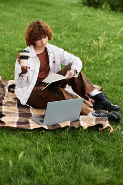 Kıvırcık saçlı genç bir kadın parktaki bir battaniyede dinlenirken dizüstü bilgisayarıyla uzaktan çalışıyor..