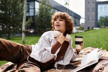 Kıvırcık saçlı genç bir kadın enerjik bir parkta battaniyede dinleniyor, kahvesini yudumluyor ve gülümsüyor..