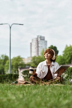 Kıvırcık saçlı kadın bir battaniyeye bağdaş kurmuş, kahvesini yudumluyor ve açık havada tablet kullanıyor..