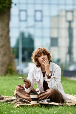 Kıvırcık saçlı genç bir kadın battaniyeye oturur, kahvesini yudumlar ve parkta doğanın tadını çıkarır..