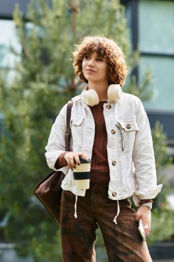 Kıvırcık saçlı genç bir kadın elinde kahve fincanıyla modern ofis binasının yanında duruyor..