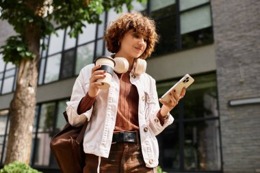 Kıvırcık saçlı sıradan giyinmiş bir kadın iş yerinin yakınında bir kahve fincanı ve akıllı telefon tutuyor..