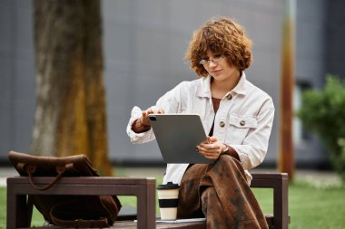 Kıvırcık saçlı kadın tabletini güneşli bir parkta kullanır ve yaz havasının tadını çıkarır..