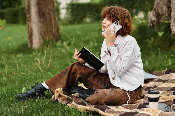 Genç bir kadın yeşil parktaki bir battaniyeye uzanıyor ve yakınlarda bir defterle telefonuyla konuşuyor..