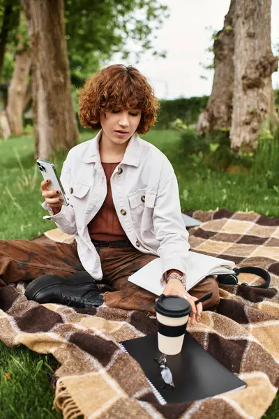 Kıvırcık saçlı genç bir kadın, güneşli bir günde battaniyeye oturup doğanın tadını çıkarıyor..