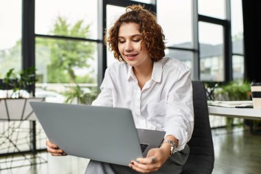 Kıvırcık saçlı iş kadını şık bir ofiste çalışırken bilgisayarına odaklanmış..