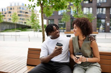 Canlı bir şehirde, genç bir çift bir bankta rahatlar içkilerini paylaşır ve birlikte gülerler..